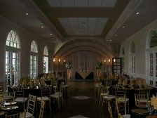 Elegant ballroom with round windows, arched ceiling, tables set for a banquet with black tablecloths and gold chairs.