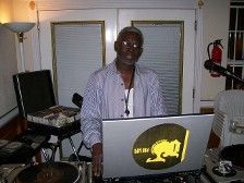 Man DJing at a party, standing in front of a laptop with a yellow logo. He wears glasses and a button-down shirt.
