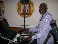 An older Black man wearing glasses and a striped shirt operates DJ equipment indoors.