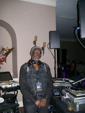 DJ with gray hair and headphones at a party, stands in front of equipment with a beige wall.