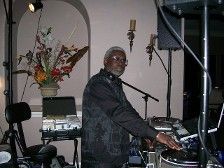 DJ at a party wearing black, using turntables and mixer. Soft lighting, floral arrangement in the background.