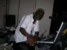 DJ at a party, working on a laptop and turntable. Wearing a white shirt, with headphones and glasses.