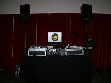 DJ setup with turntables, laptop, speakers, and a black table against a red curtain.