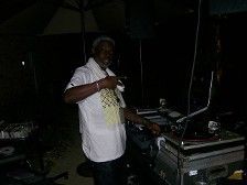 DJ in white shirt at a dark outdoor event, pointing, near equipment.