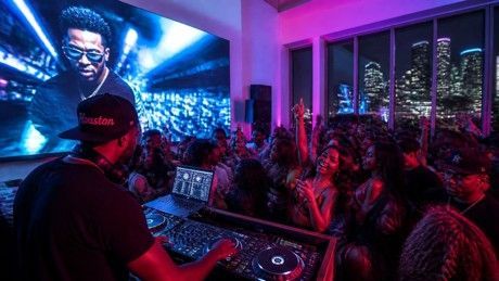 Crowded neon-lit club dance party with DJ on stage and people dancing and cheering