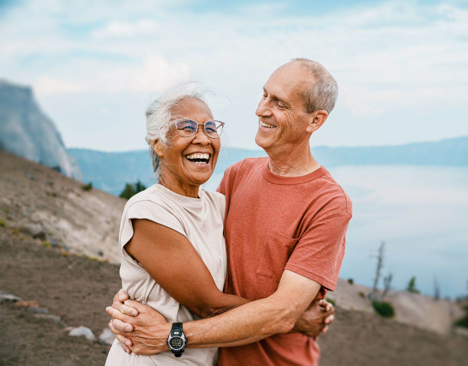An elderly couple embracing and laughing outdoors, with a mountain and lake in the background.