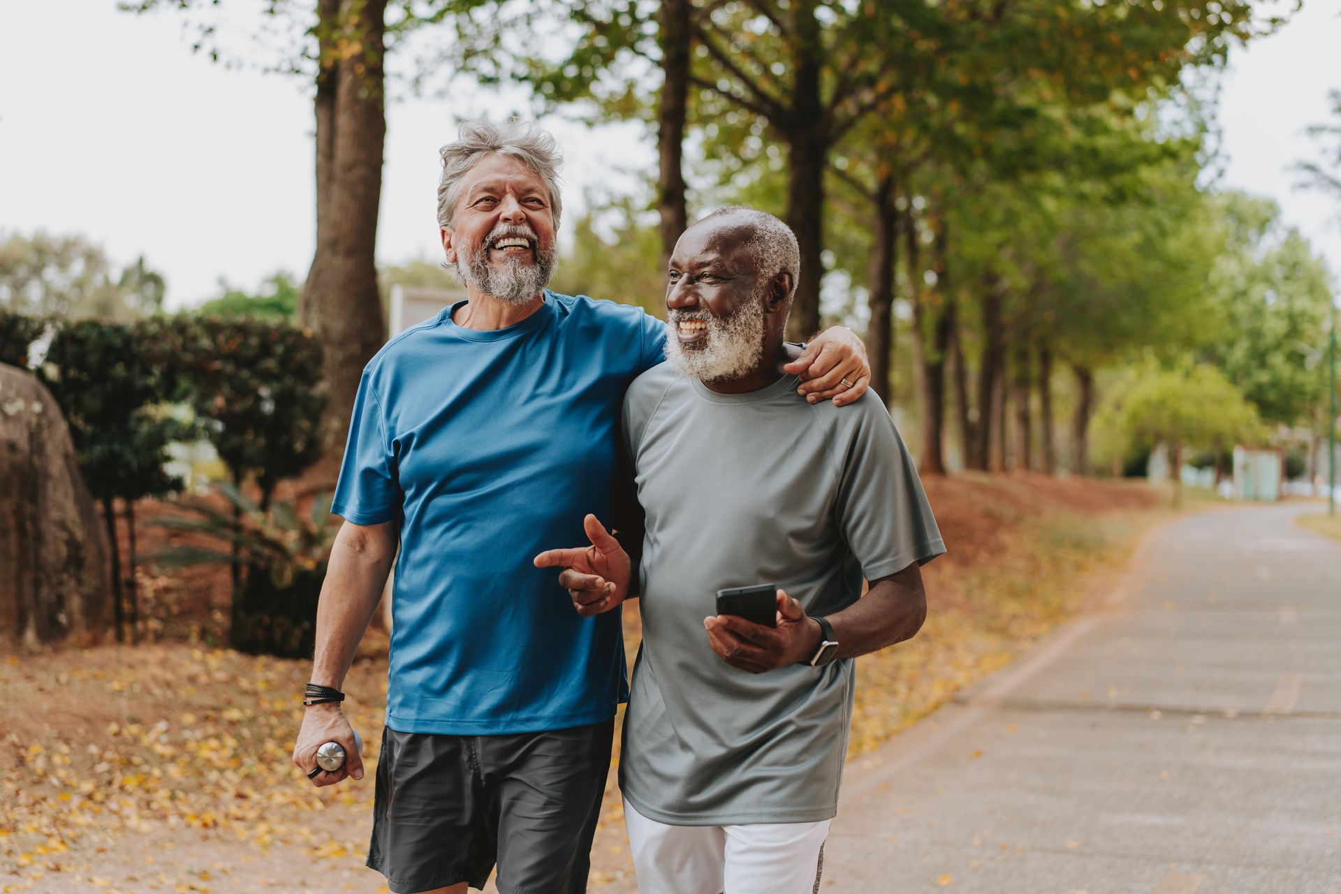 Two older men walking together on a path, one arm around the other. Trees line the path.