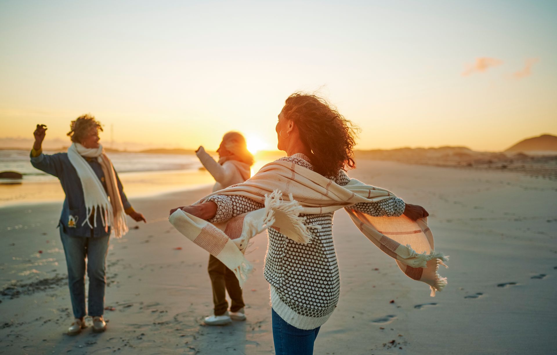 Three women dancing on a beach at sunset; the sky is orange, and they wear sweaters and scarves.
