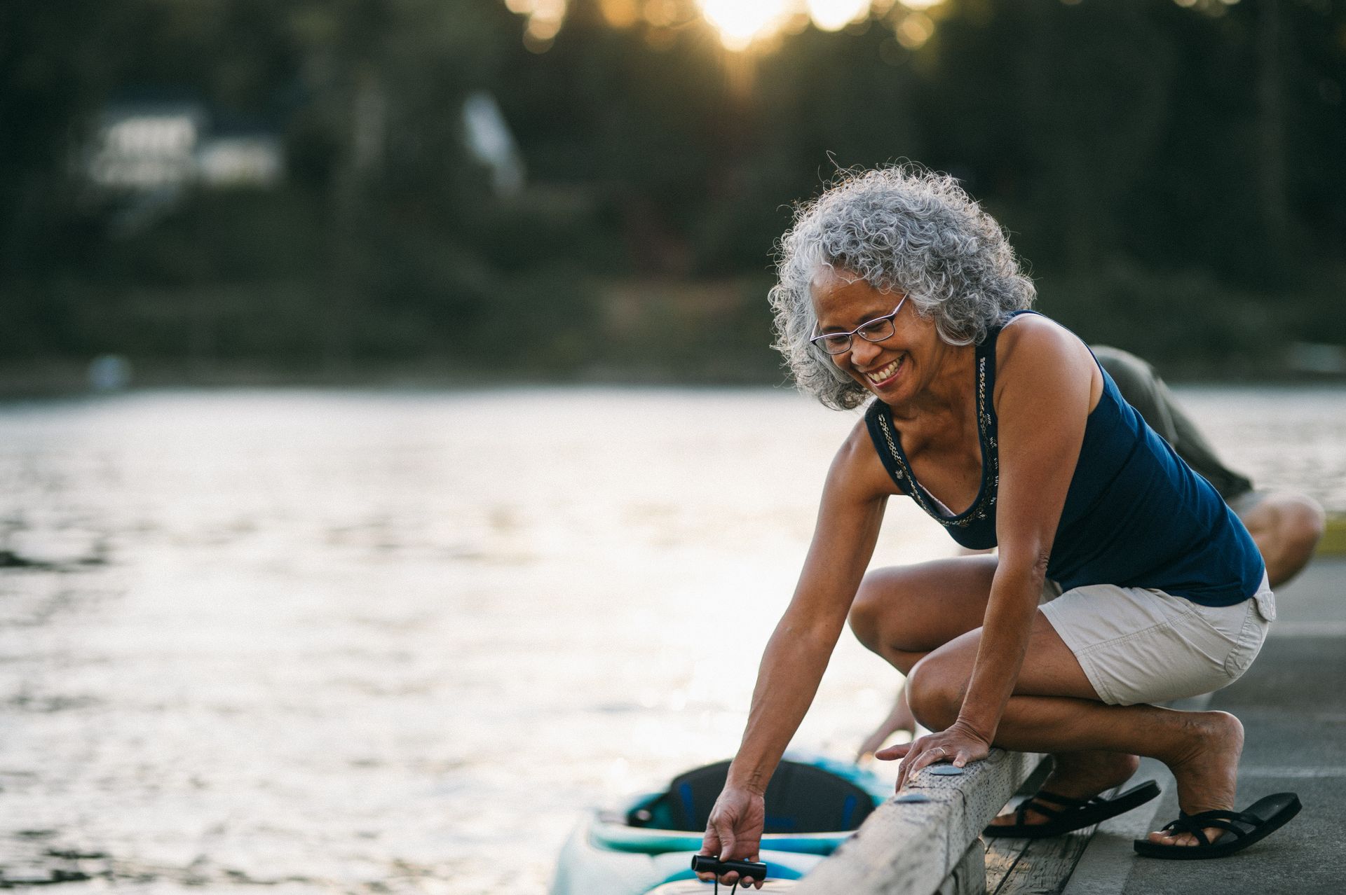 Woman in shorts and tank top smiles as she crouches near a kayak at the water's edge.