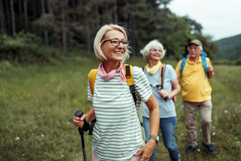 Three smiling older adults hike through a grassy field with trees.