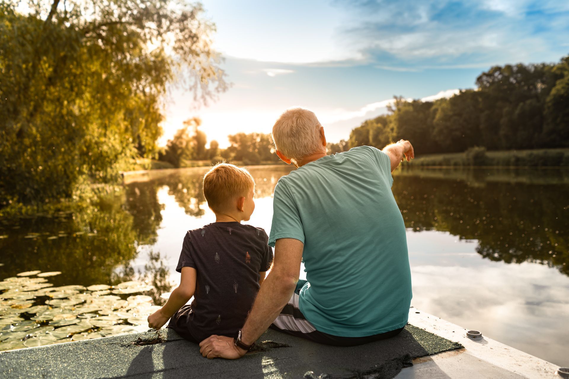 Man and boy sit on dock, looking at a lake at sunset. Man points, both silhouetted, with trees and water reflecting the sun.