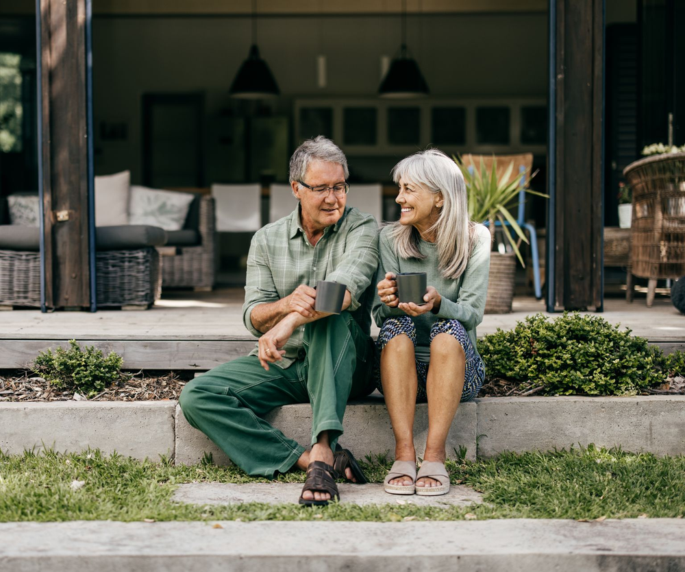 Couple sitting on steps, smiling, holding mugs, in front of a house.