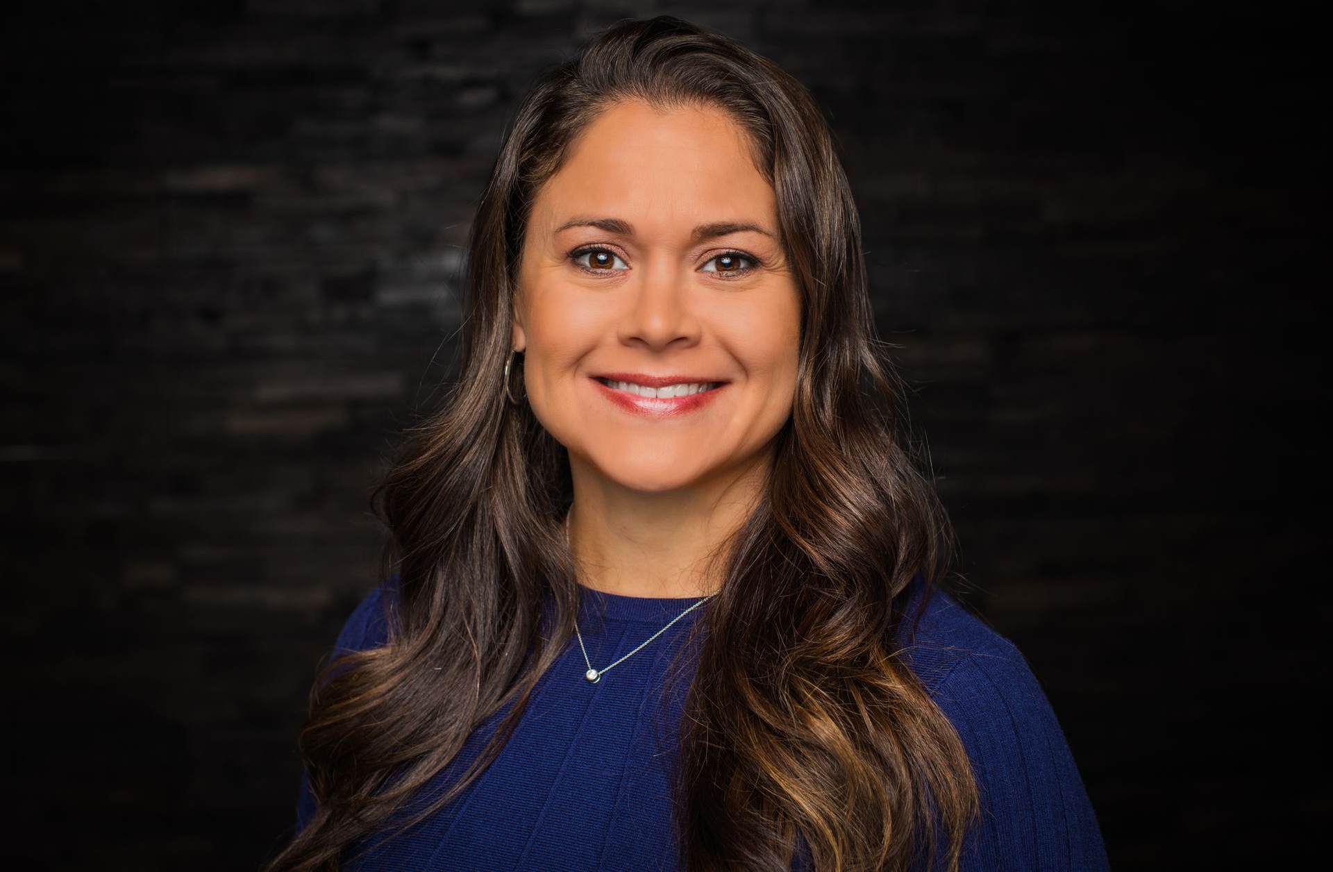 Woman with long brown hair smiles; navy blue sweater; black background.