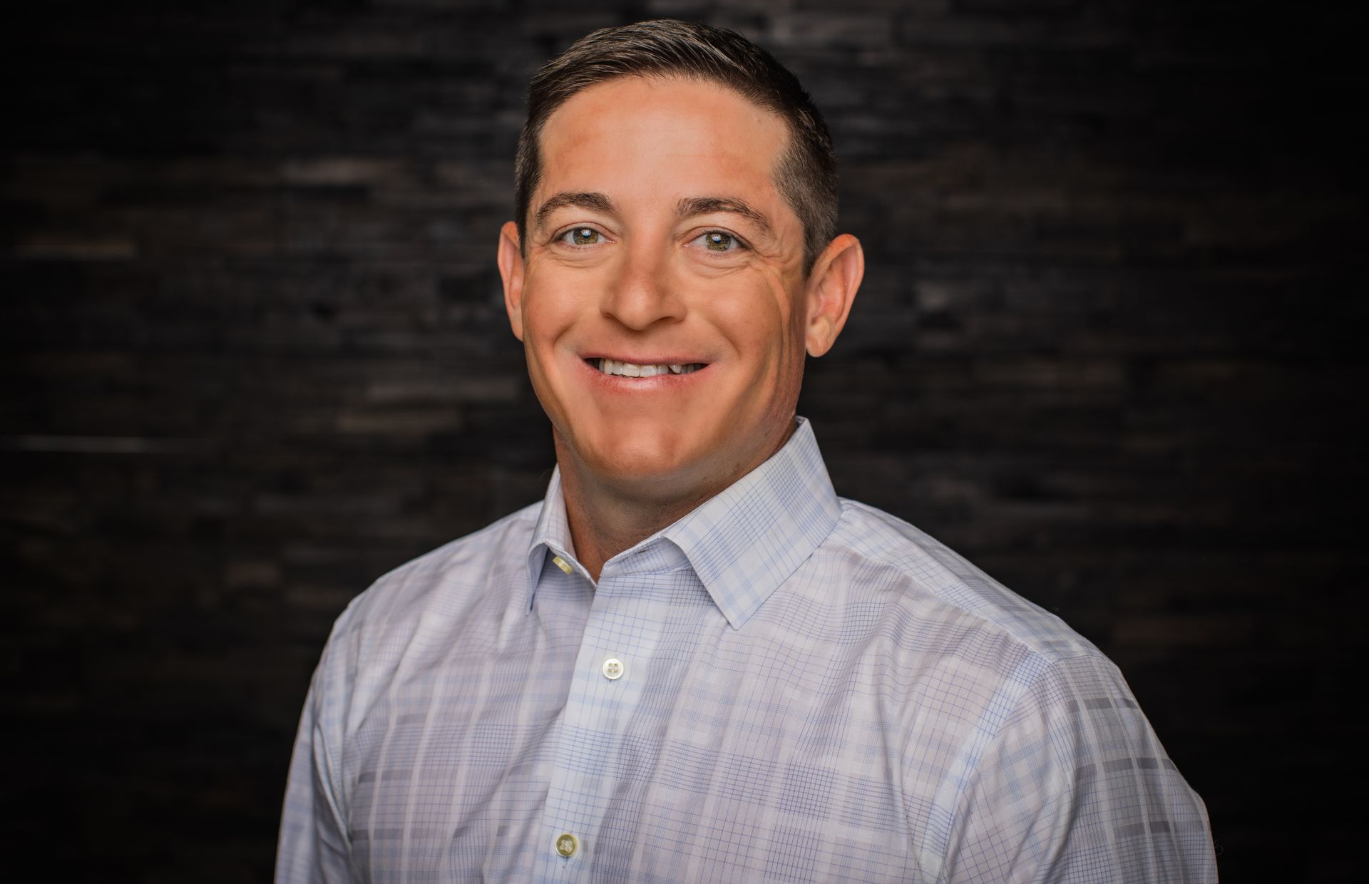 Man in a light blue collared shirt smiles, dark wood background.