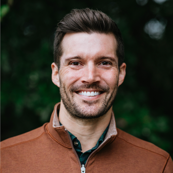 A man with a beard is smiling for the camera while wearing a brown jacket.