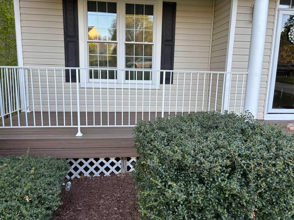 Front porch with white railing, beige siding, window, and trimmed hedges bordering a brick walkway.