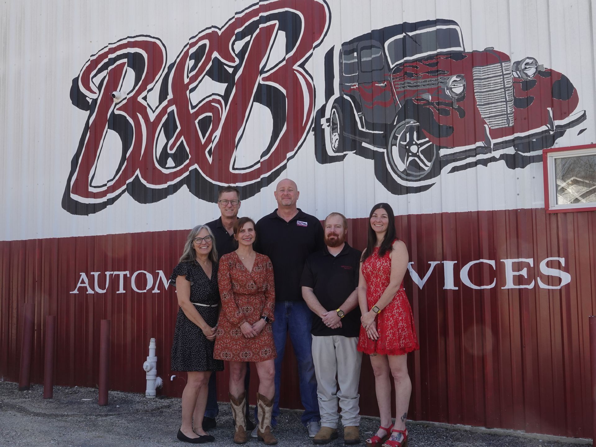 Group posing in front of B&B Automotive Services sign and vintage red car mural