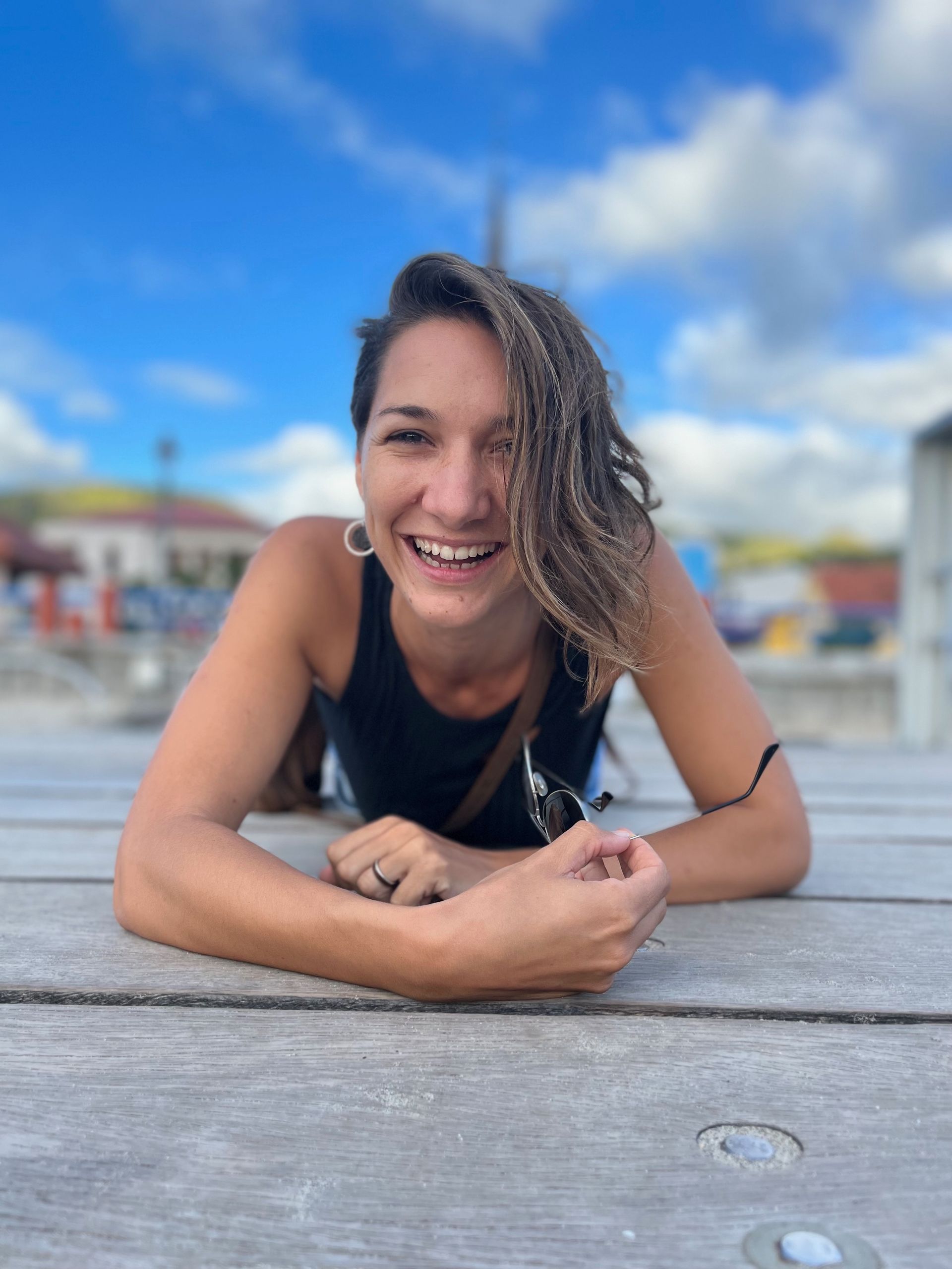 Une personne souriante, appuyée sur une table en bois à l'extérieur, sous un ciel bleu nuageux.