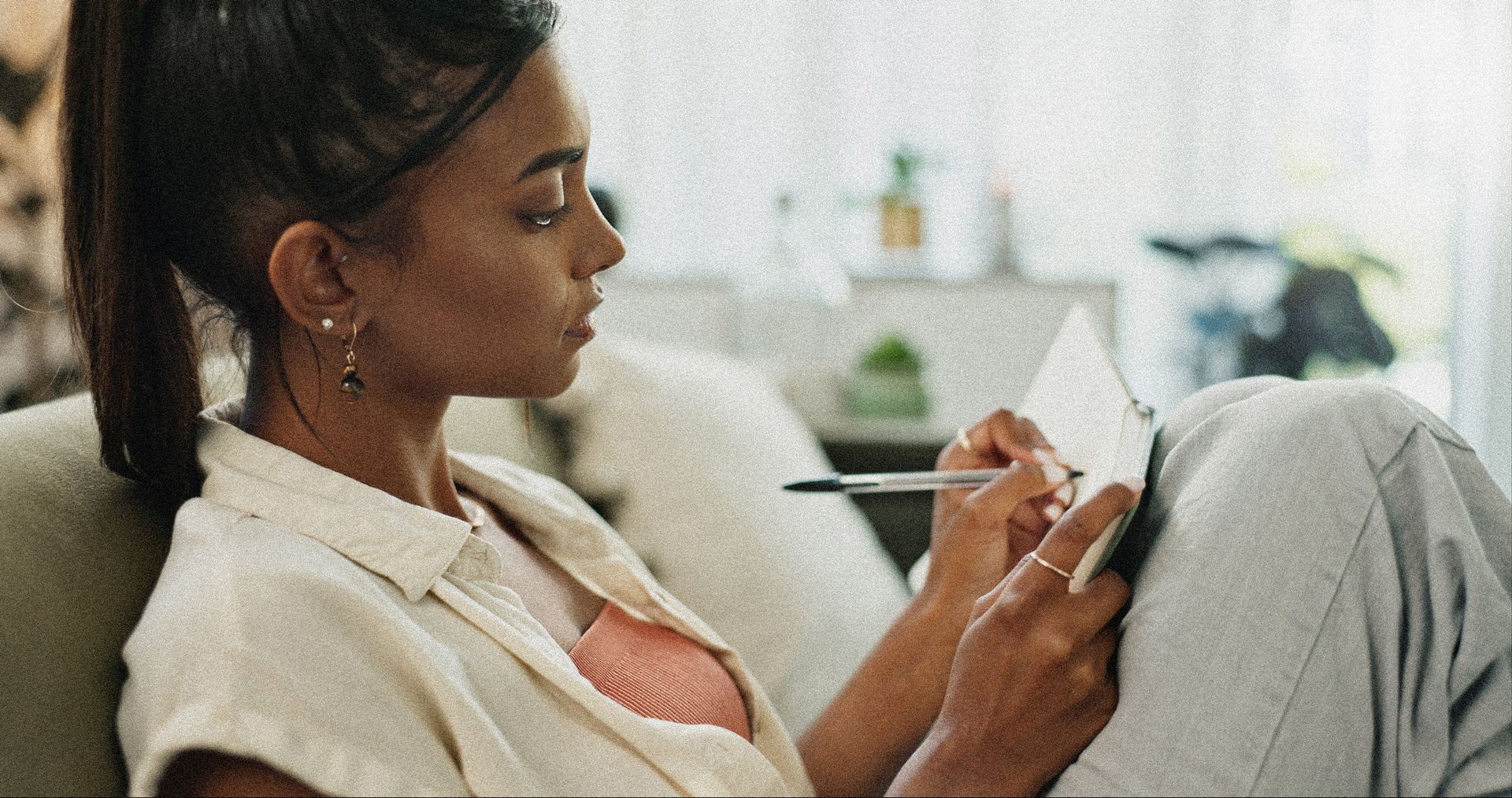 Femme allongée sur un canapé, tenant un téléphone et un stylo dans un salon lumineux