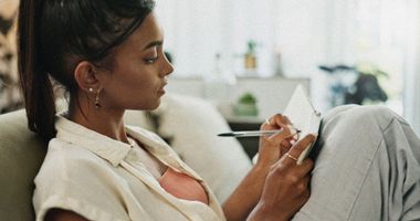 Une femme allongée sur un canapé regarde son téléphone à côté d'un carnet et d'un stylo.