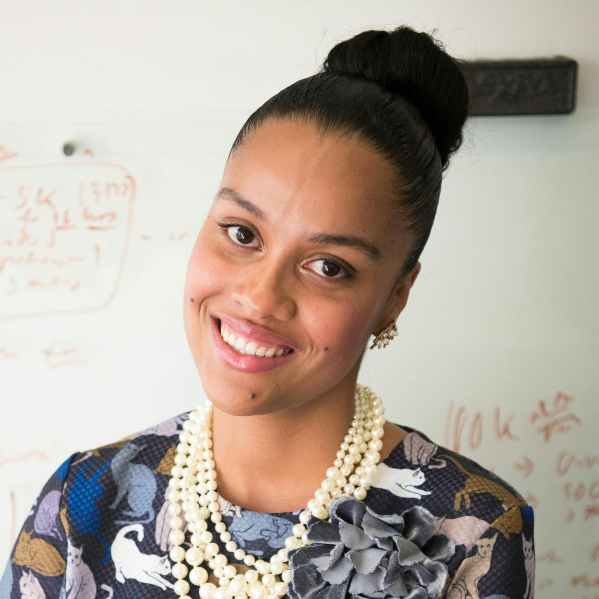 Femme souriante en robe à fleurs et collier de perles, debout devant un tableau blanc.