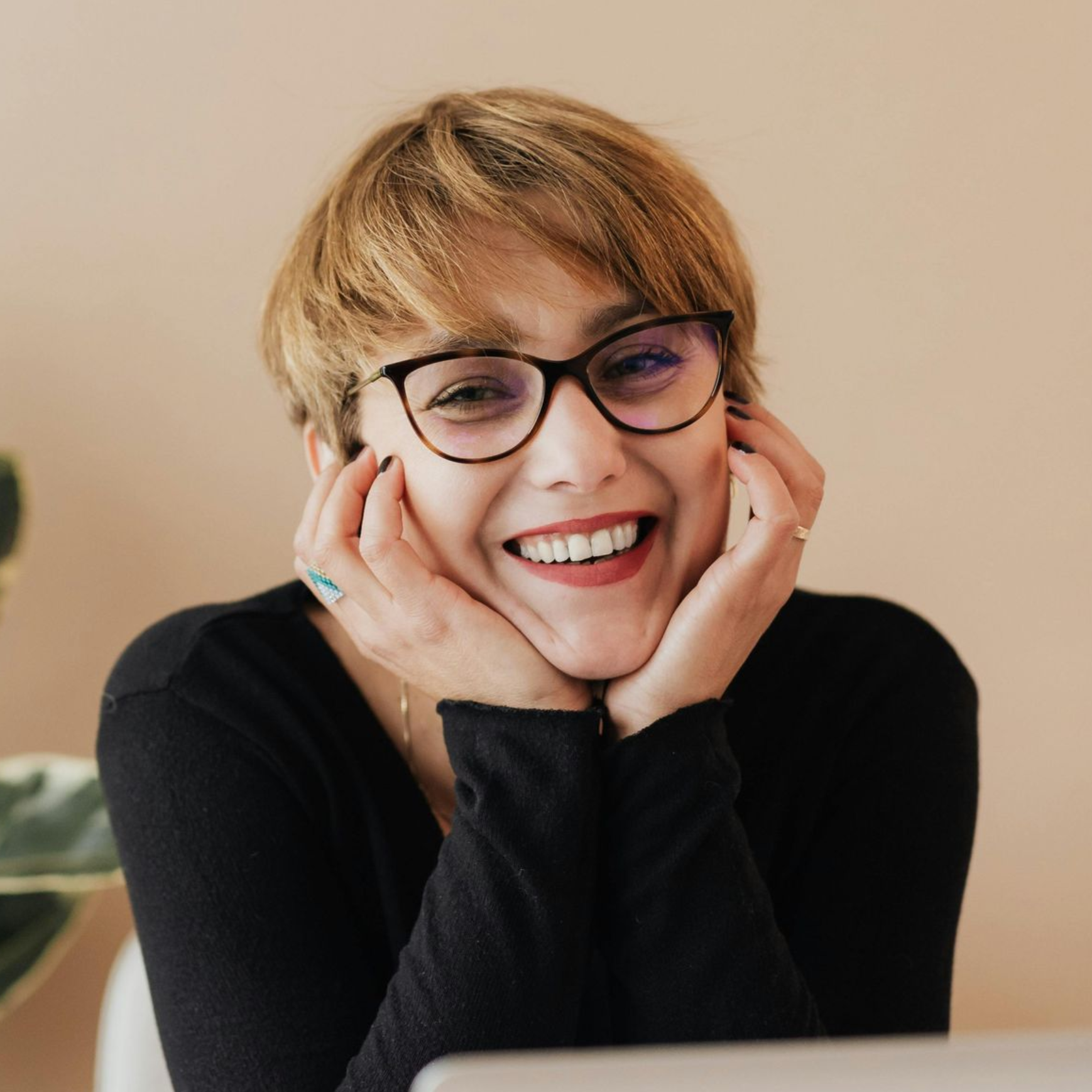 Personne souriante portant des lunettes, le visage appuyé sur ses mains, sur fond beige