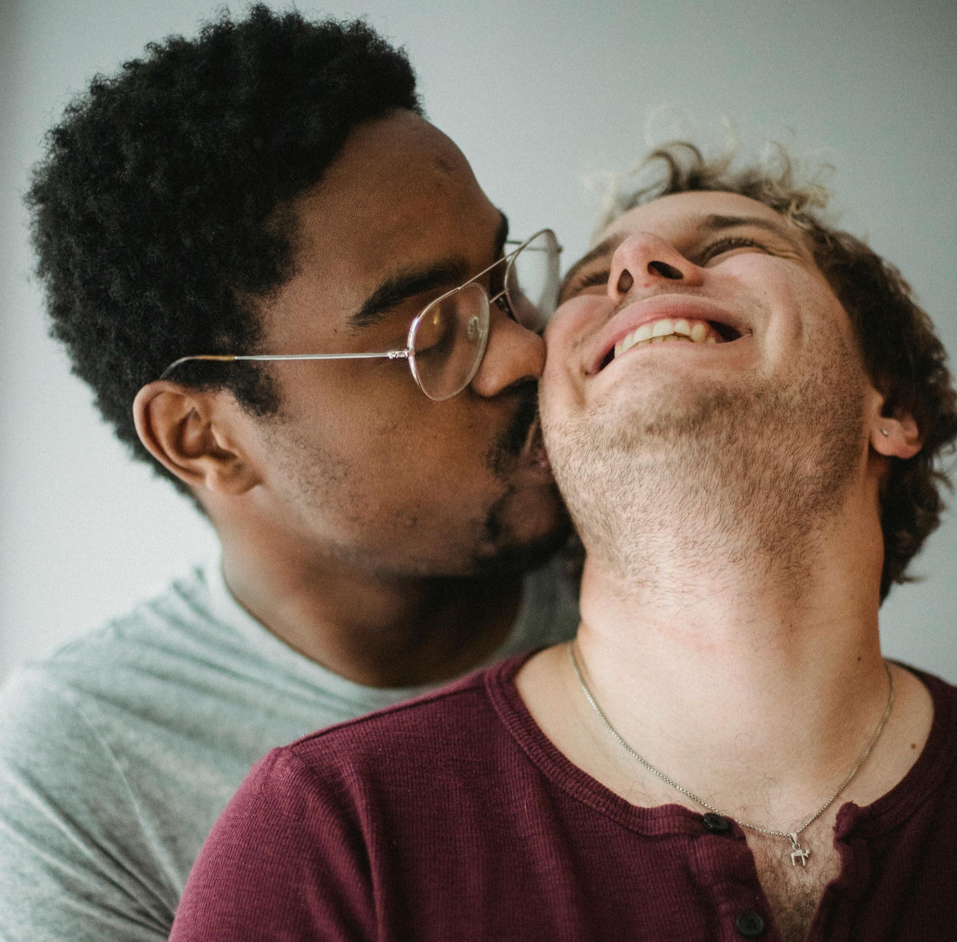 Deux personnes proches l'une de l'autre, l'une embrassant la joue de l'autre tandis qu'elles sourient devant un mur nu.