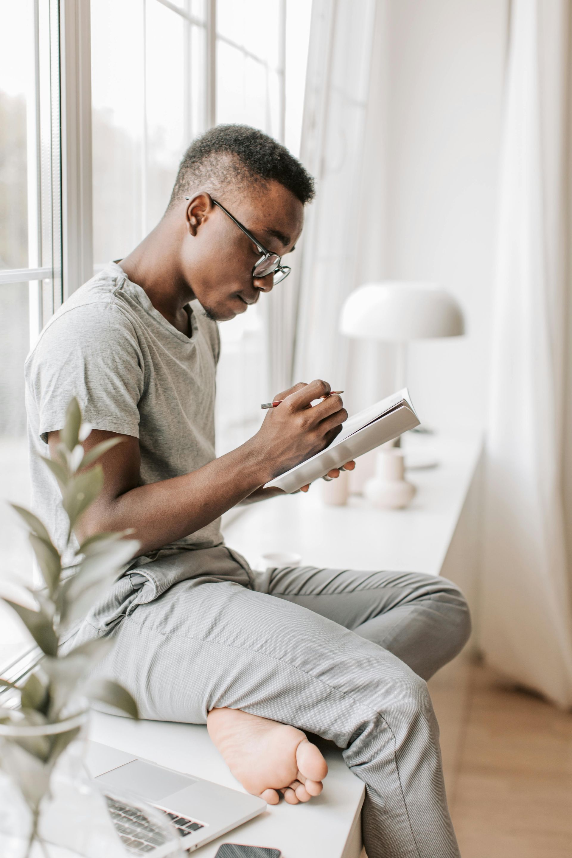 Une personne assise près d'une fenêtre, écrivant dans un carnet sur un lit avec une lampe à proximité.