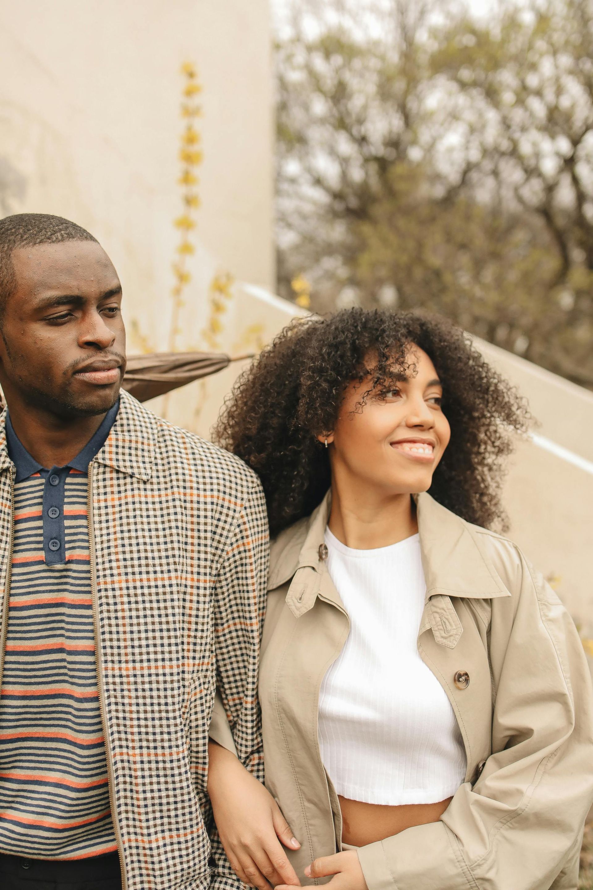 Deux personnes marchent dehors, l'une d'elles, vêtue d'un trench-coat beige, sourit, près d'un mur couleur crème et d'arbres.