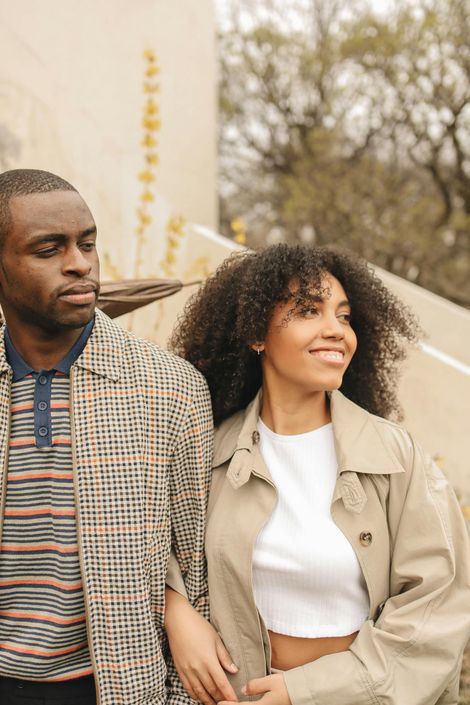 Deux personnes marchent dehors, l'une d'elles, vêtue d'un trench-coat beige, sourit, près d'un mur couleur crème et d'arbres.