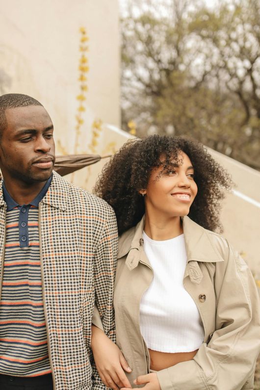 Deux personnes marchent dehors, l'une d'elles, vêtue d'un trench-coat beige, sourit, près d'un mur couleur crème et d'arbres.