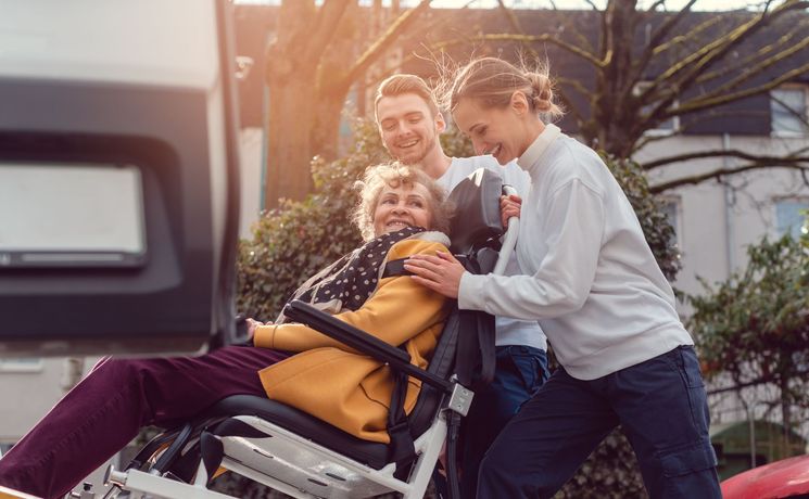 Woman in wheelchair with two people, smiling, near a vehicle.