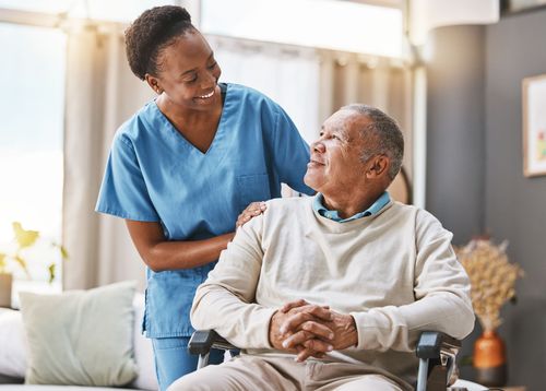 Caregiver in blue scrubs smiles at a man in a wheelchair, indoors.
