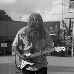A man with dreadlocks is holding a guitar in a black and white photo.