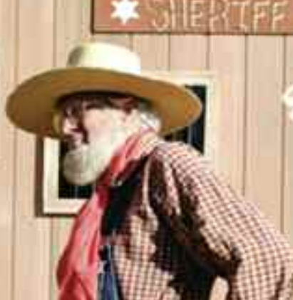 A man with a beard and a hat is standing in front of a sheriff 's sign.