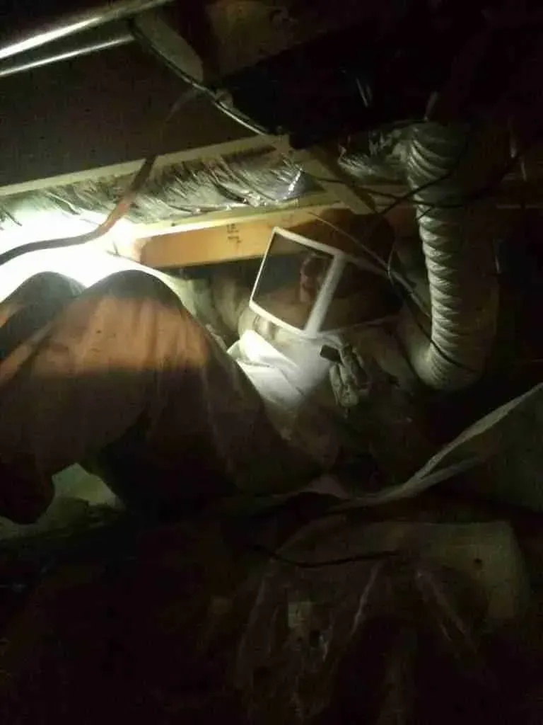 Person in protective suit working under a house, near ductwork and insulation.