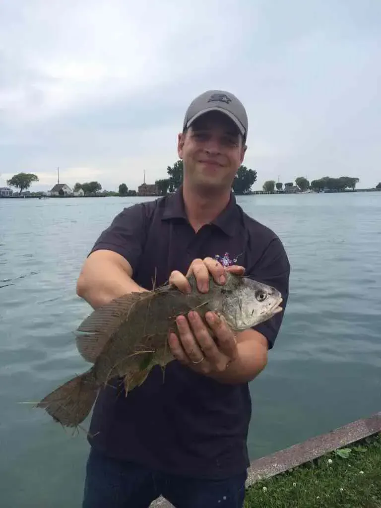 Man holding a fish by a body of water on a cloudy day.