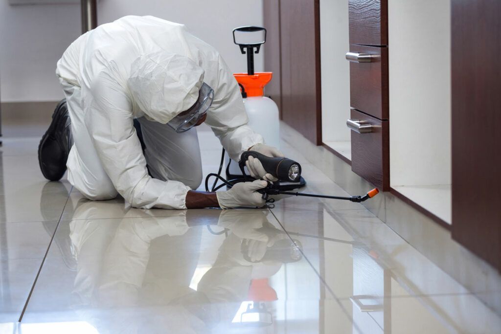 Pest control technician kneeling, spraying baseboard, wearing protective suit, in room with cabinets.