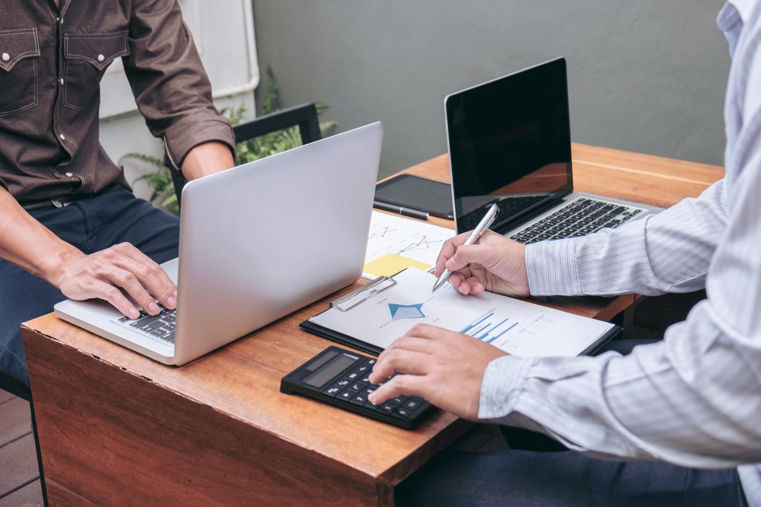 Two people at a desk with laptops, documents, and a calculator, collaborating outdoors.