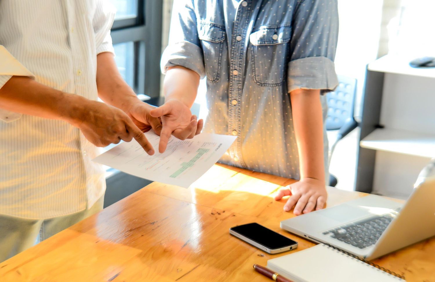 Two people pointing at a document on a wooden desk with a laptop, phone, and notebook.