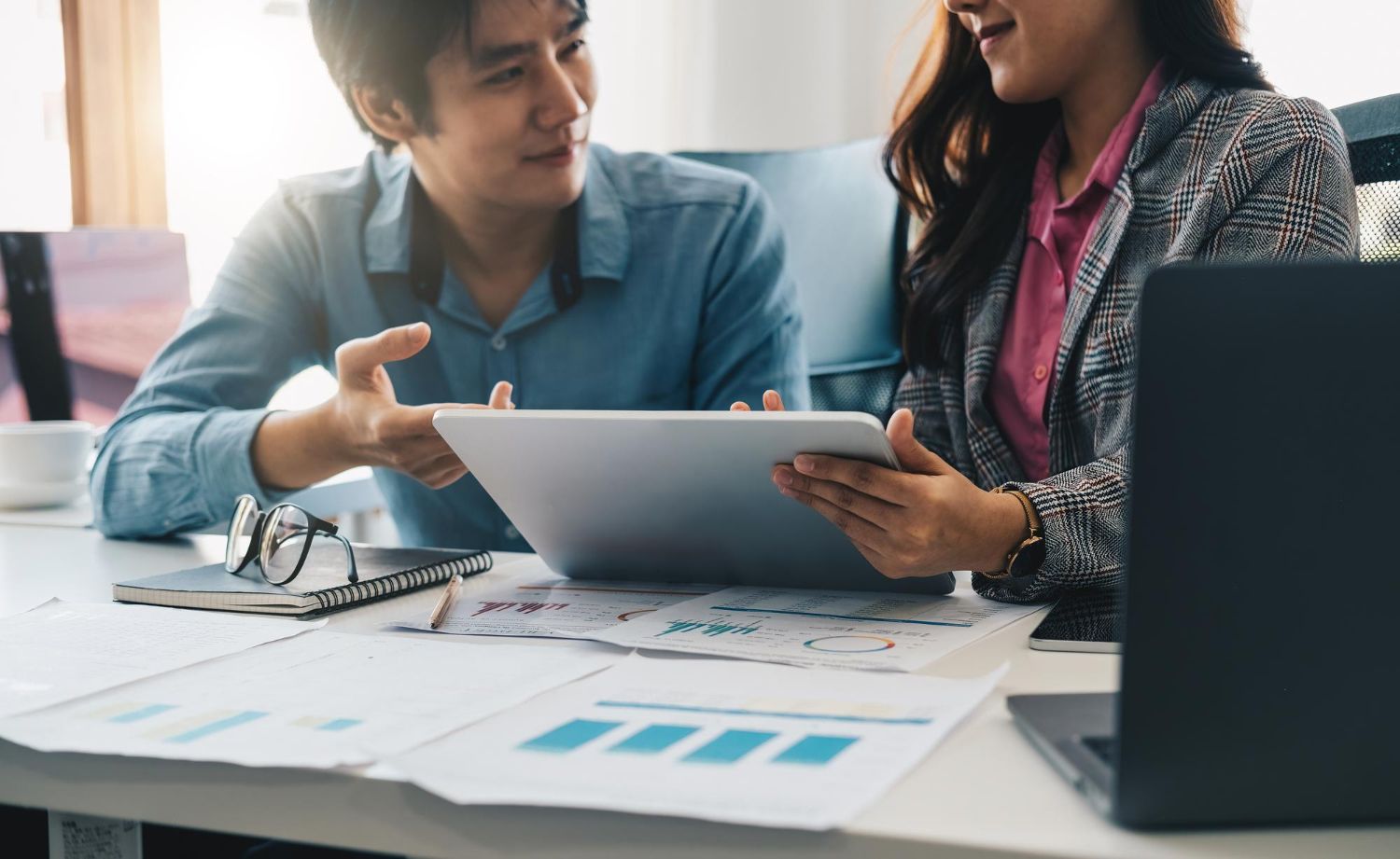 Two people reviewing data on a tablet at a desk with charts and a laptop.