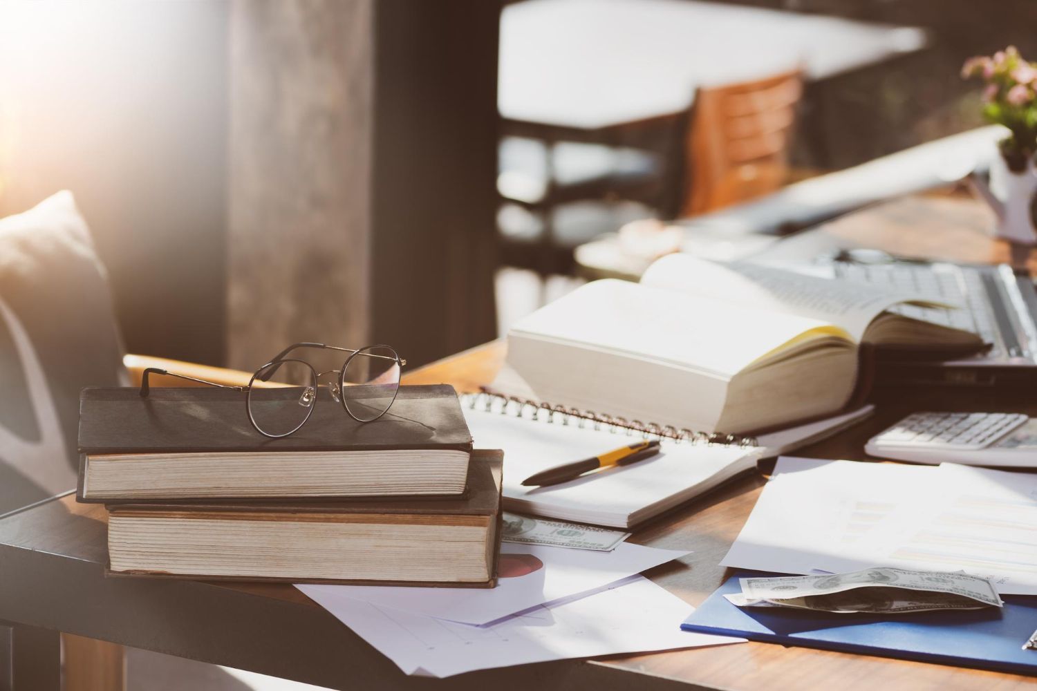 Books, notebook, laptop, and eyeglasses on a wooden table, illuminated by sunlight.