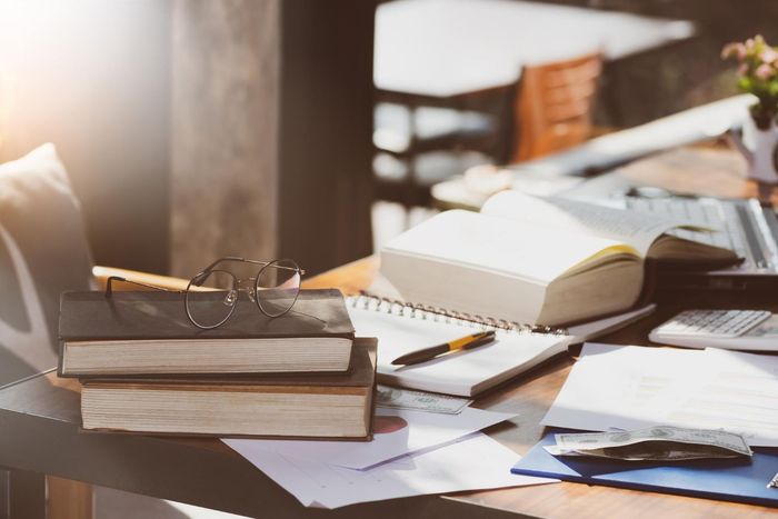 Books, notebook, laptop, and eyeglasses on a wooden table, illuminated by sunlight.