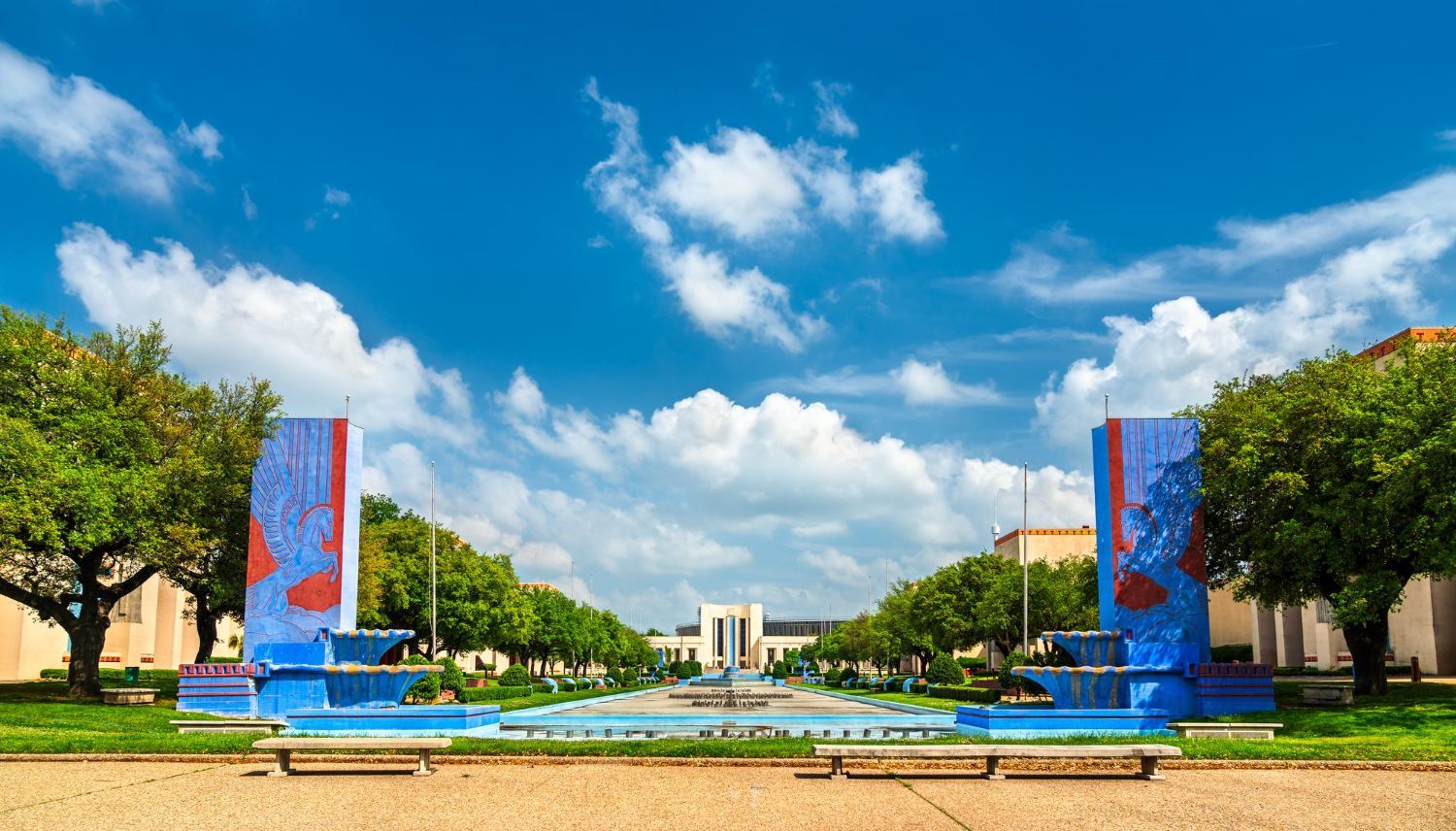 Fountain and colorful sculptures framing a path leading to a building under a blue sky with fluffy clouds.