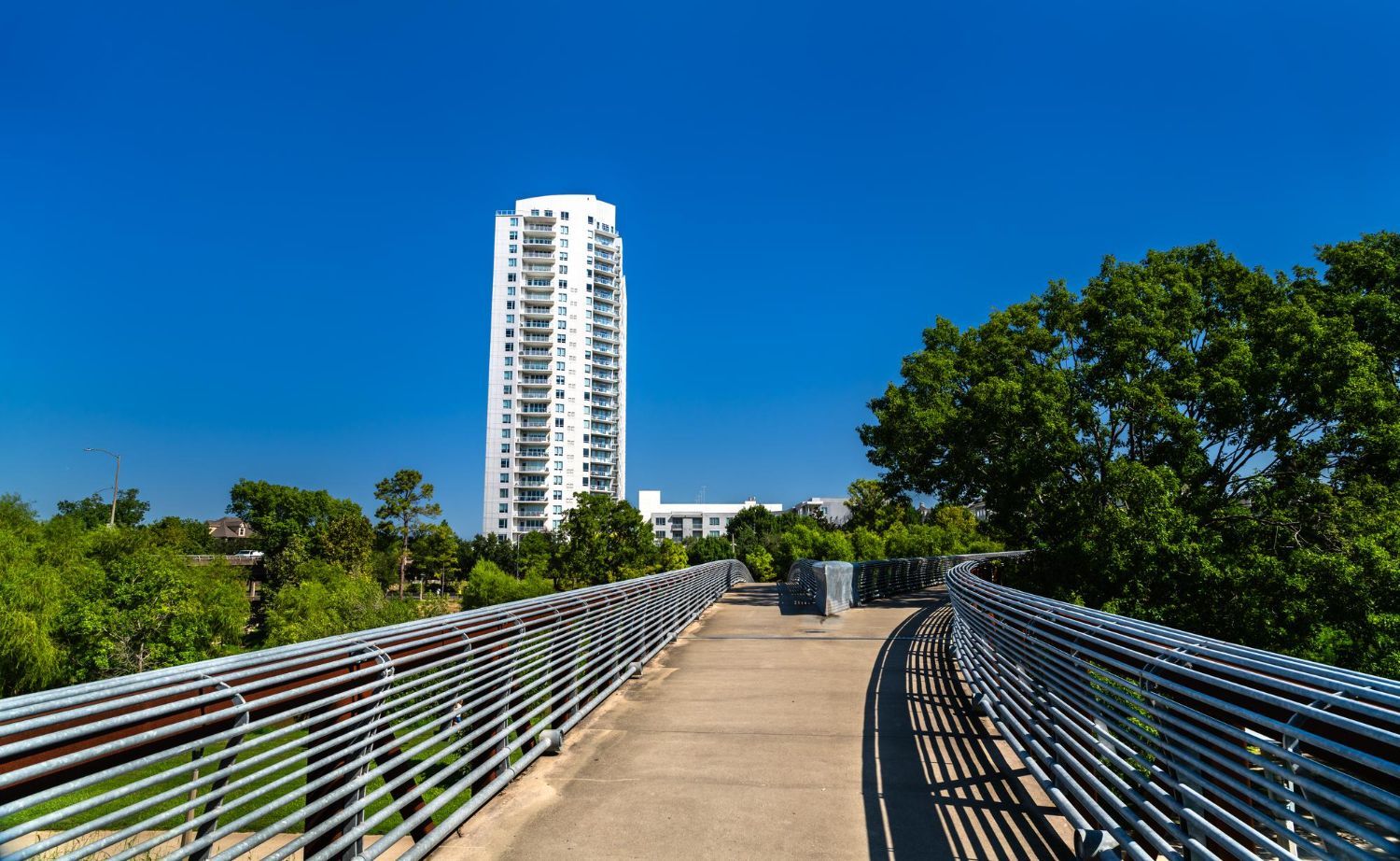 Elevated walkway with metal railings, leading to a tall white high-rise building on a bright blue day.