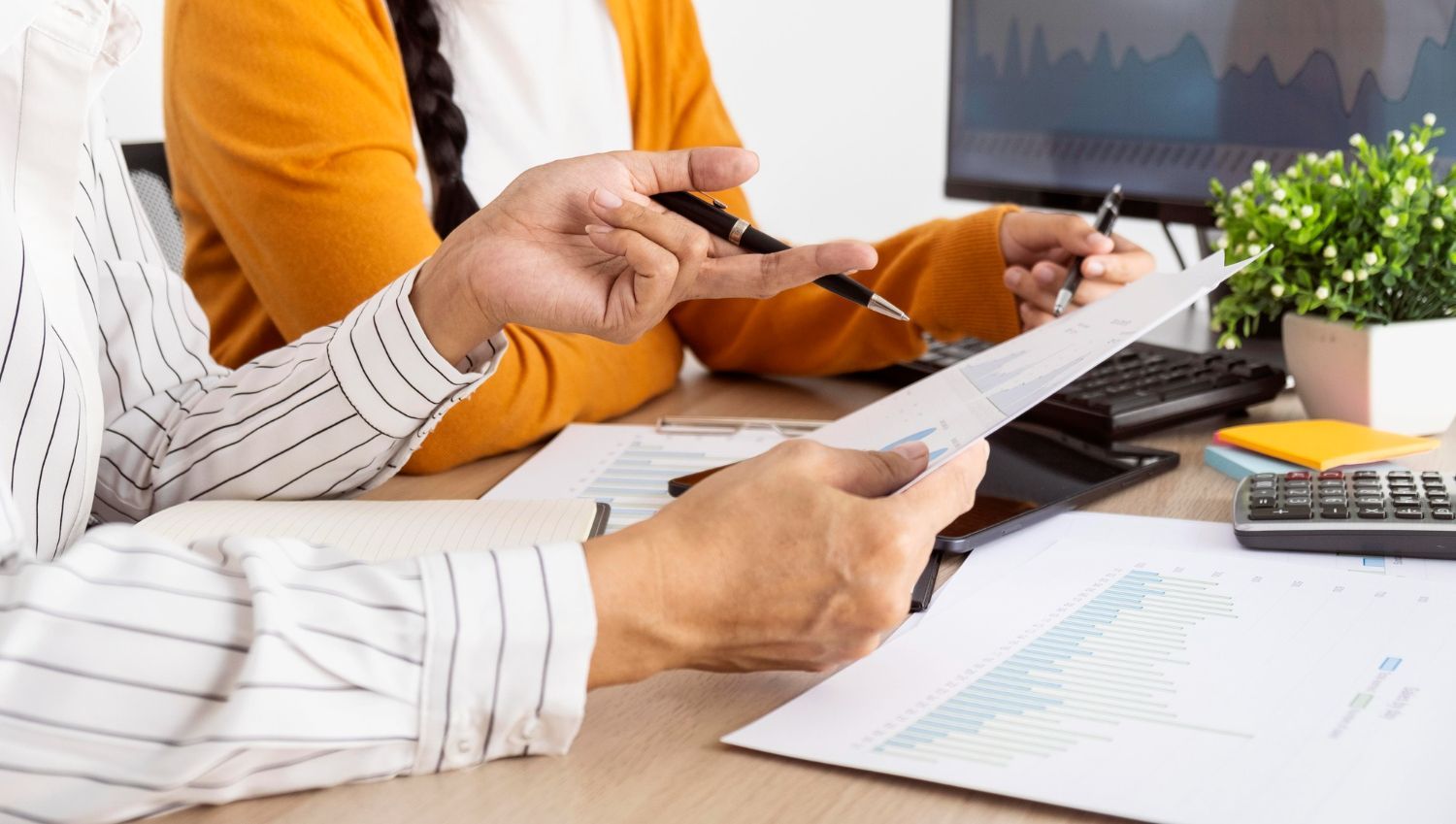 Two people at a desk review papers; one points with a pen, another holds a document, with graphs and a calculator visible.