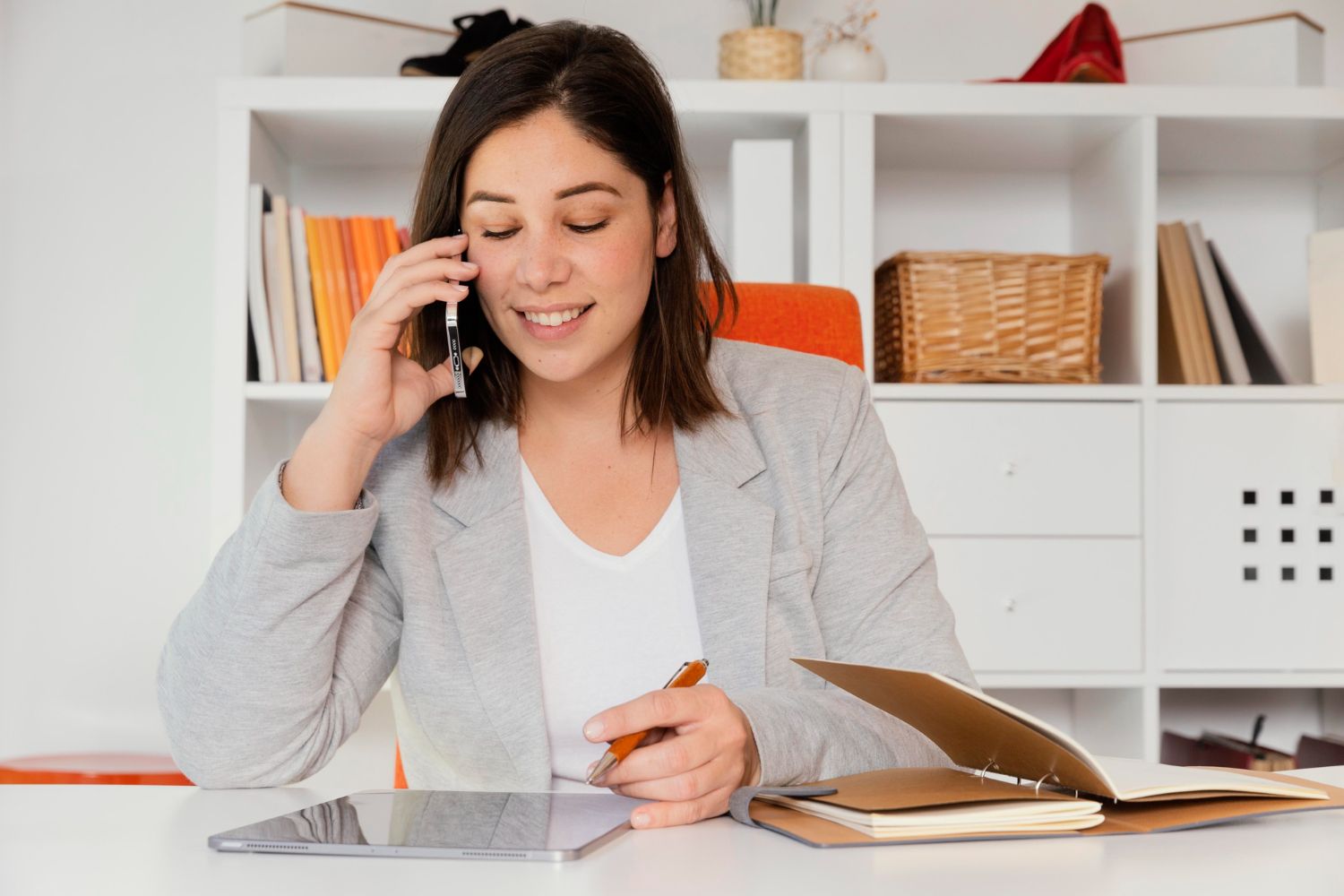 Woman smiling while talking on phone, taking notes at desk in office setting.