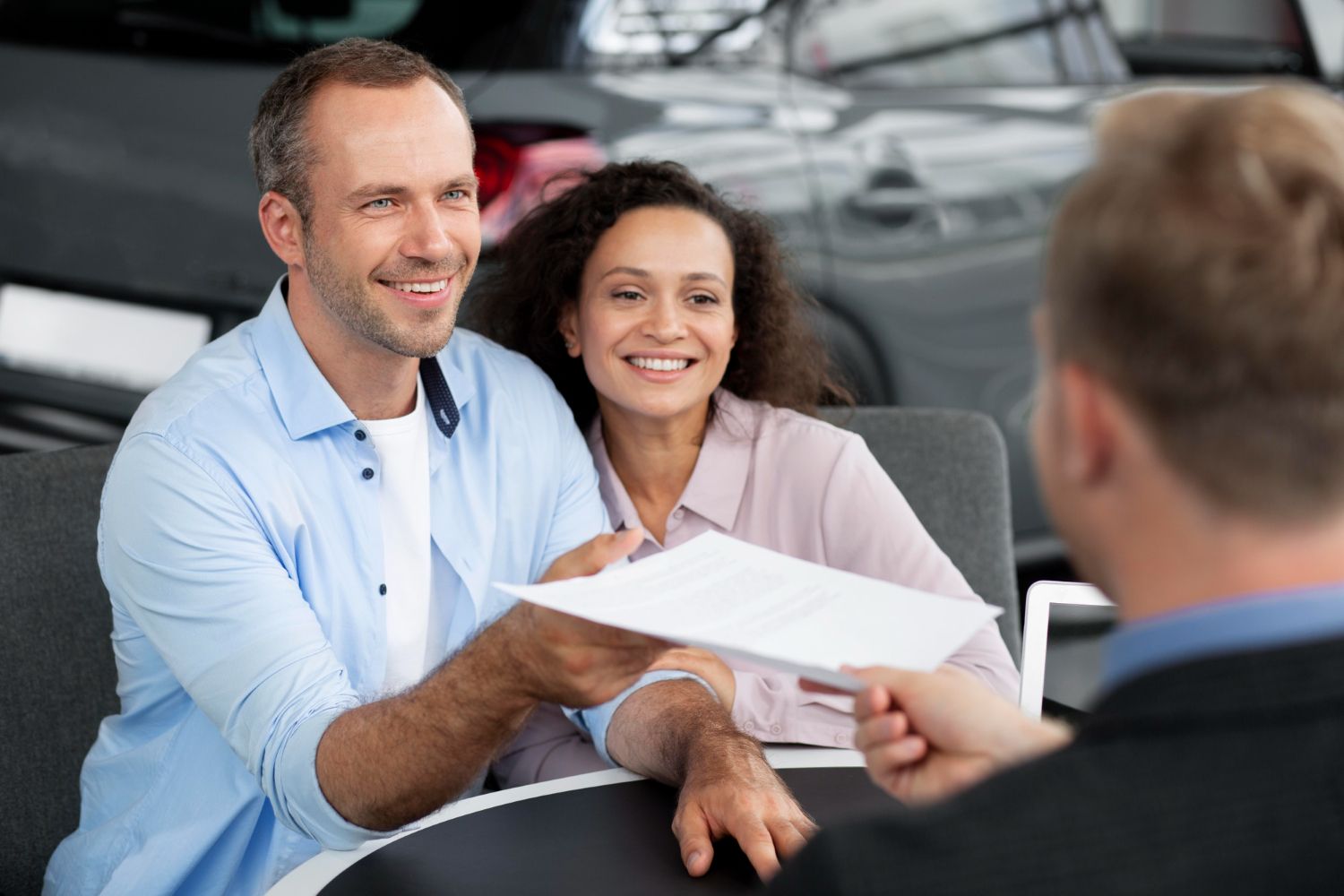Couple smiles as a salesperson hands them paperwork in a car dealership.