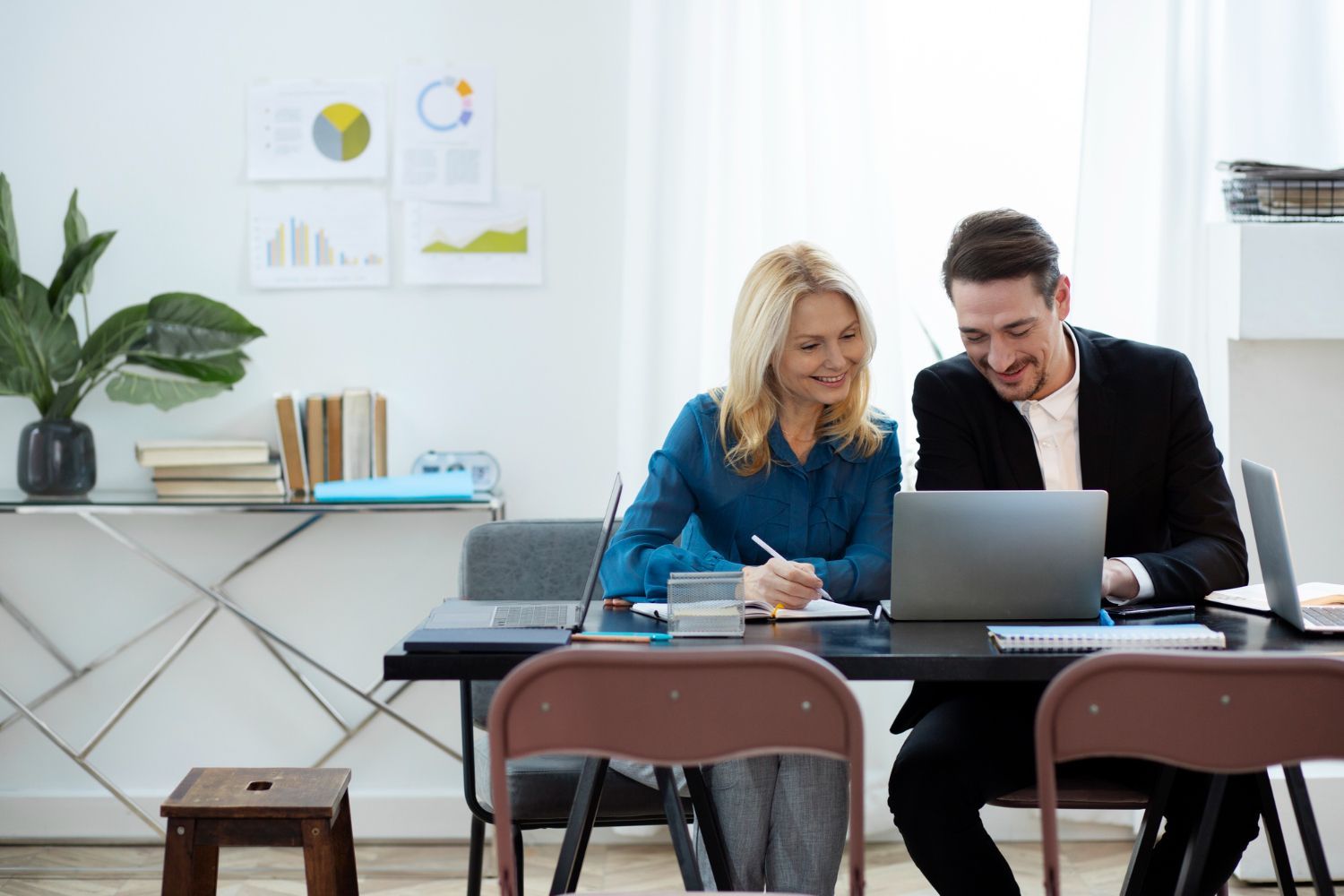 Woman and man smiling, working together at a desk on a laptop. Charts on the wall.