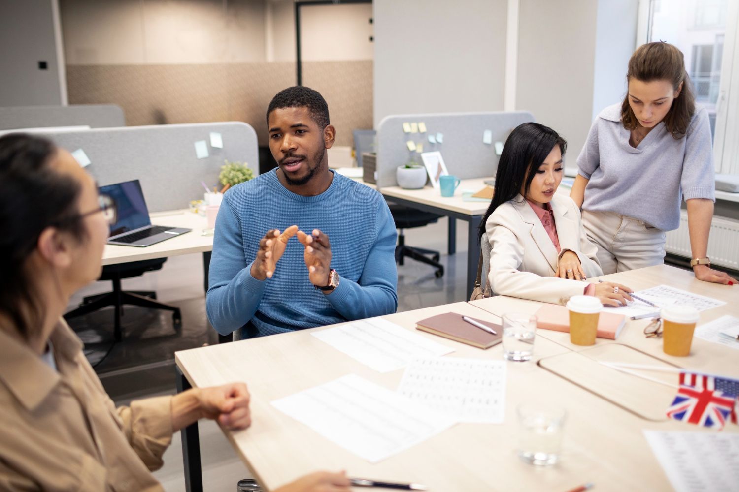 People in an office meeting around a table, one man gesturing with hands, English flag.